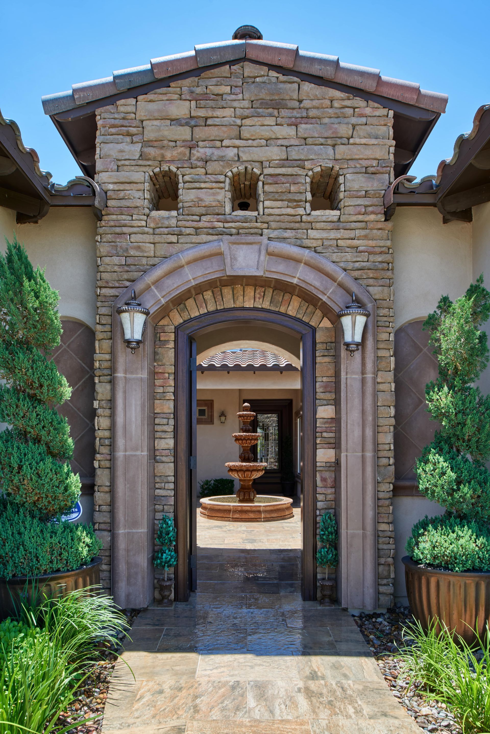 Stone archway entrance to a courtyard with a fountain, flanked by potted trees.