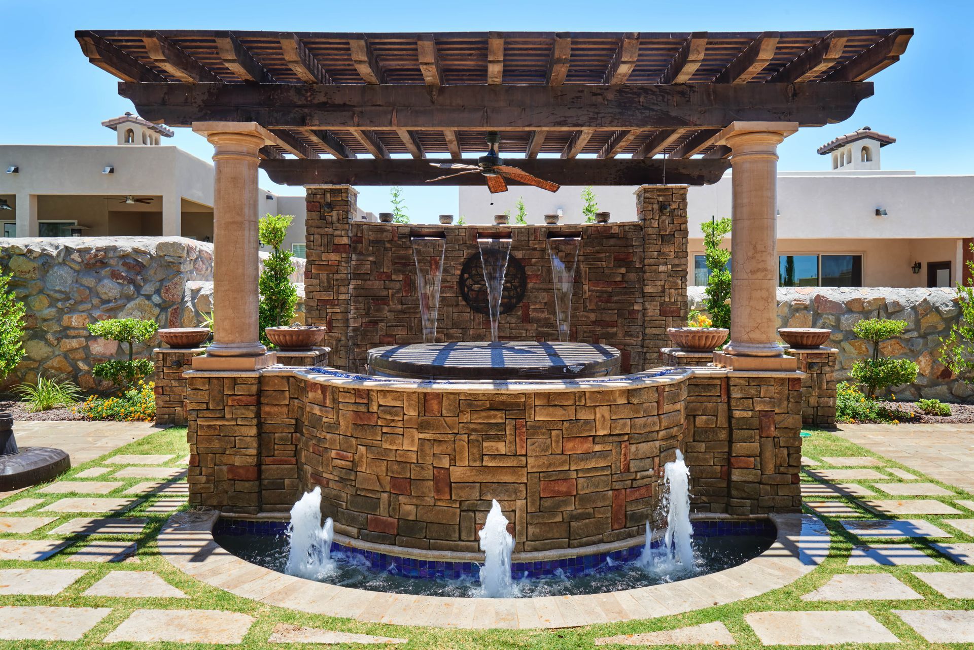 Stone fountain with water features beneath a wooden pergola; located outdoors in a garden setting.