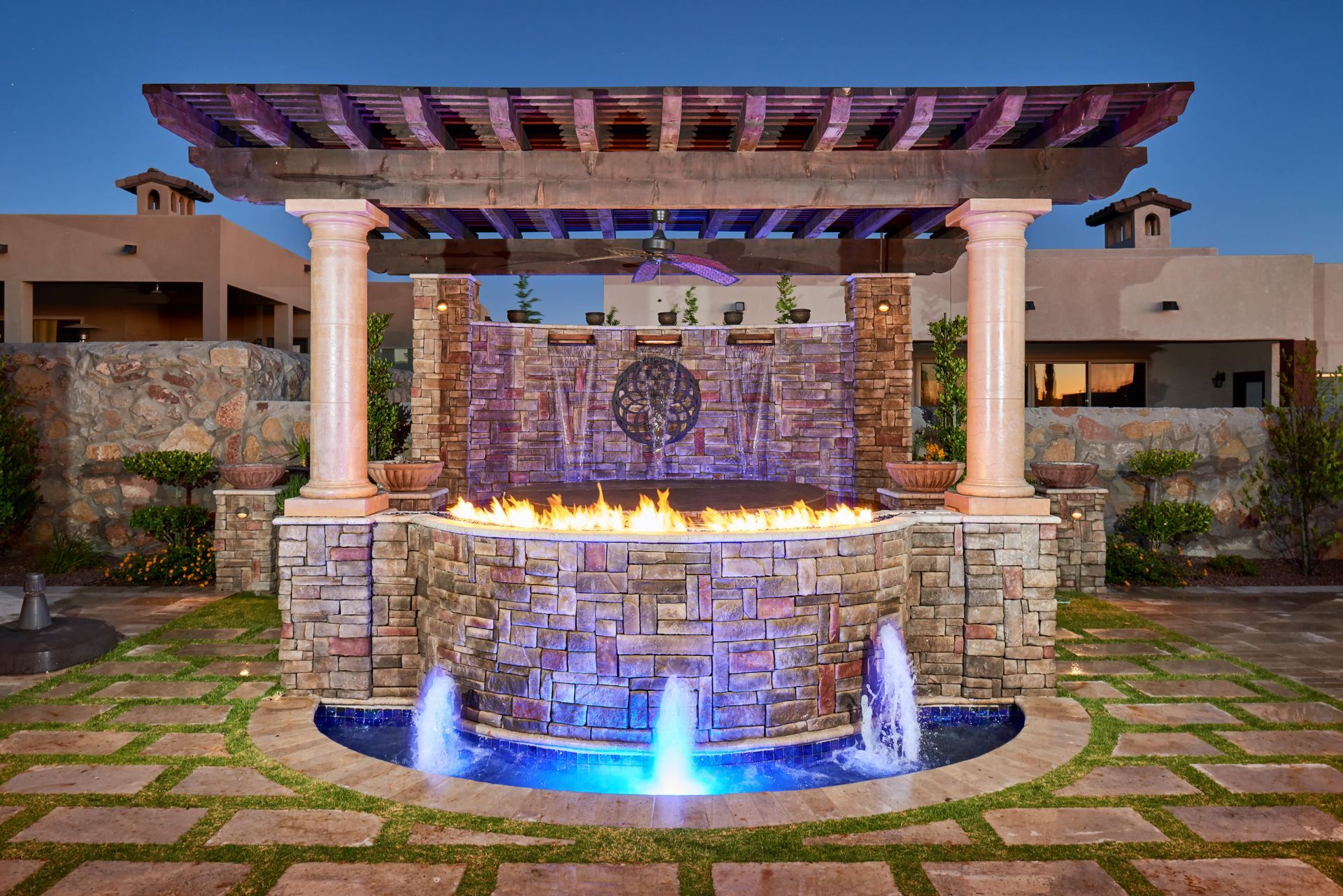 Stone water fountain with pergola and fire feature; blue and orange lighting.