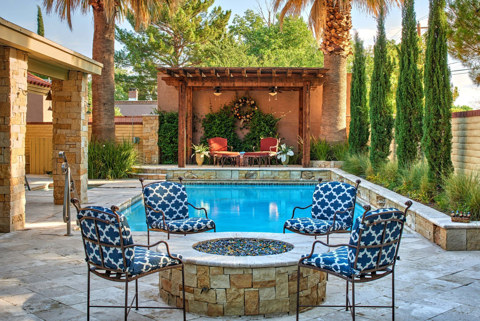 Poolside patio with fire pit, blue chairs, pergola, and seating area. Palm trees and greenery.