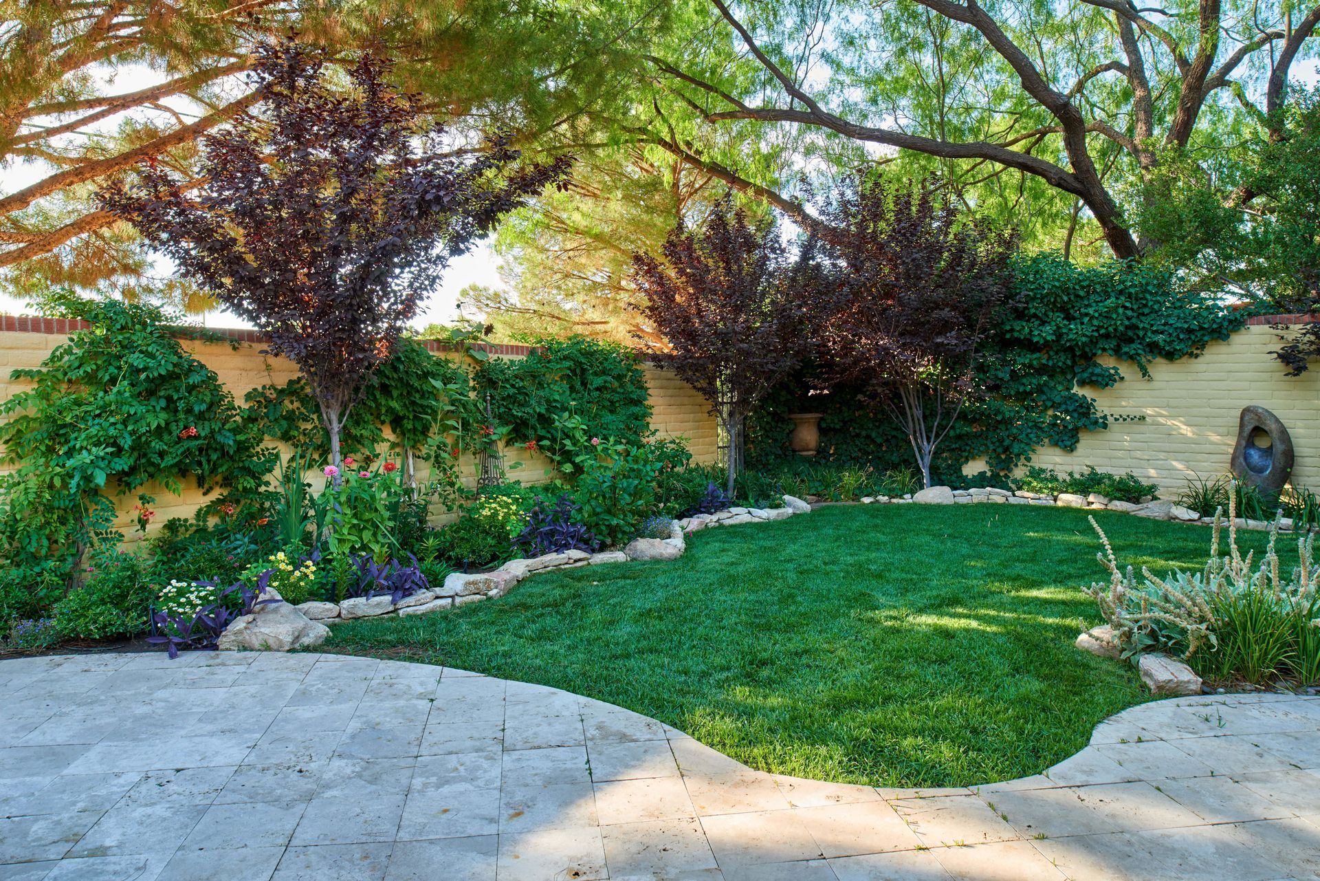 A lush, green backyard with stone patio, lawn, and flowering trees against a wall.