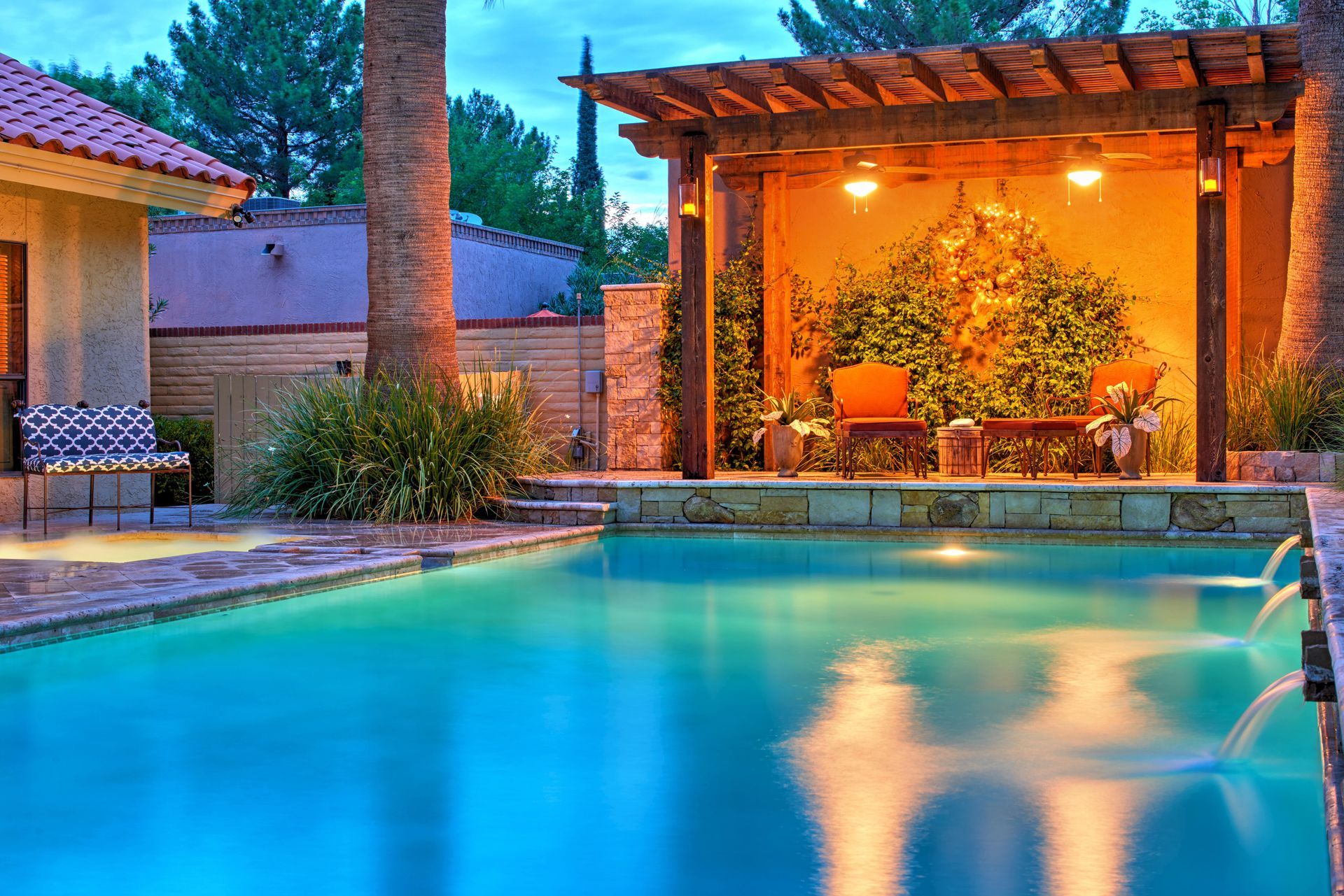Swimming pool with illuminated pergola, lounge furniture, and water fountains.