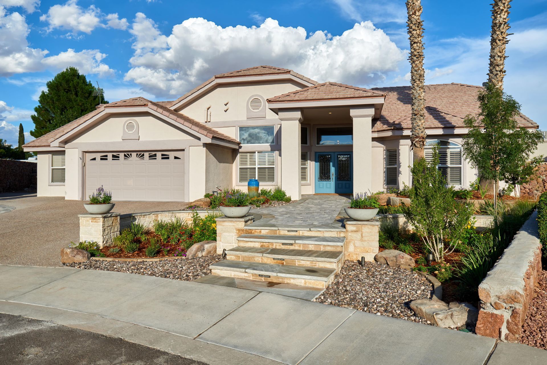 Beige house with a blue door, stone steps, and palm trees. Sunny day with clouds.