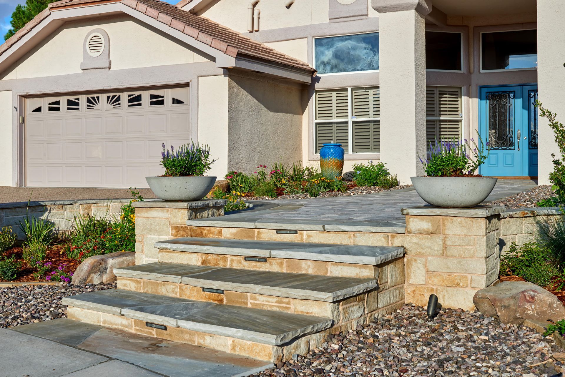 Stone steps lead to a house with a blue door, two planters, and a tan garage.