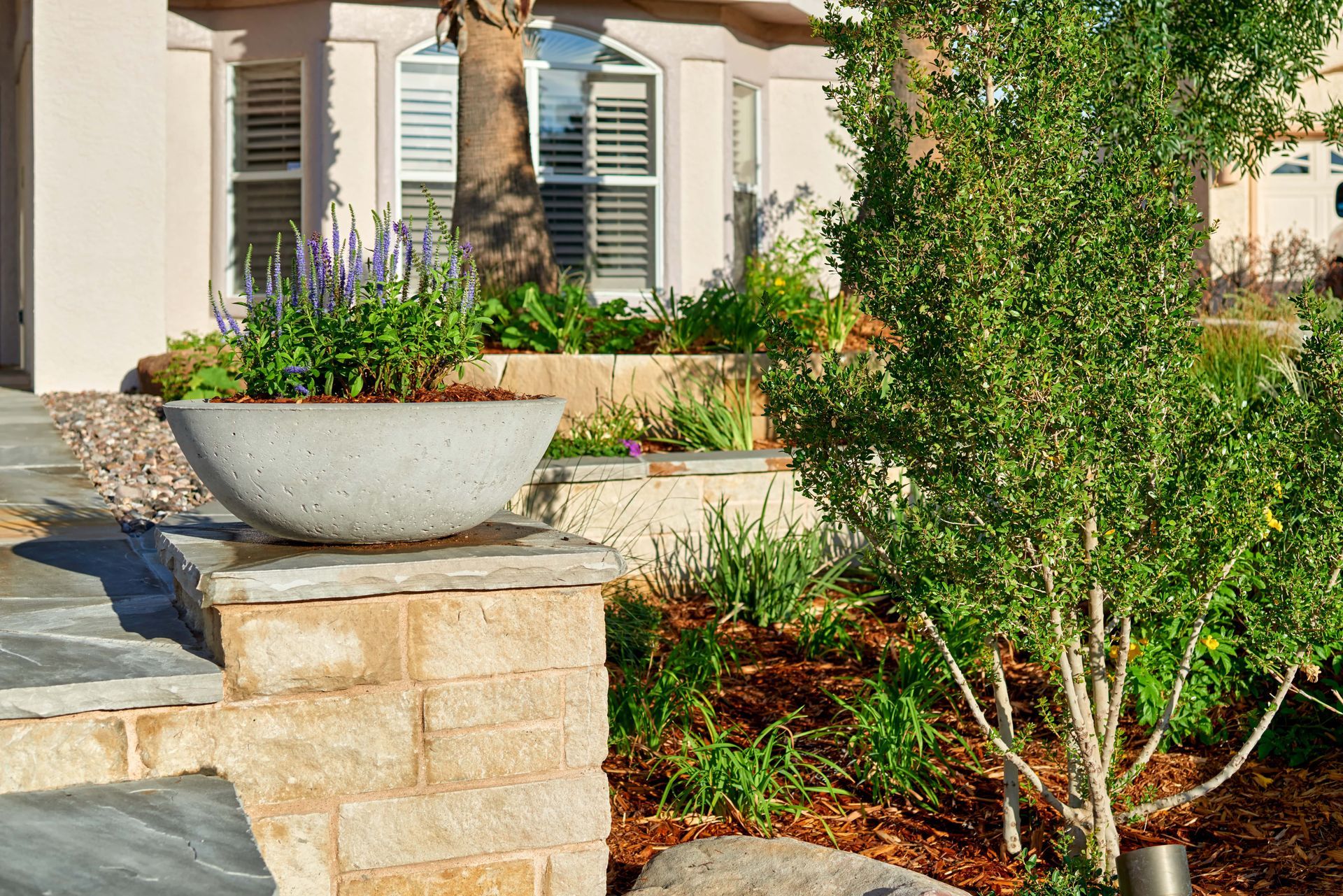 Large stone planter with lavender flowers on a brick pillar, near landscaped garden.