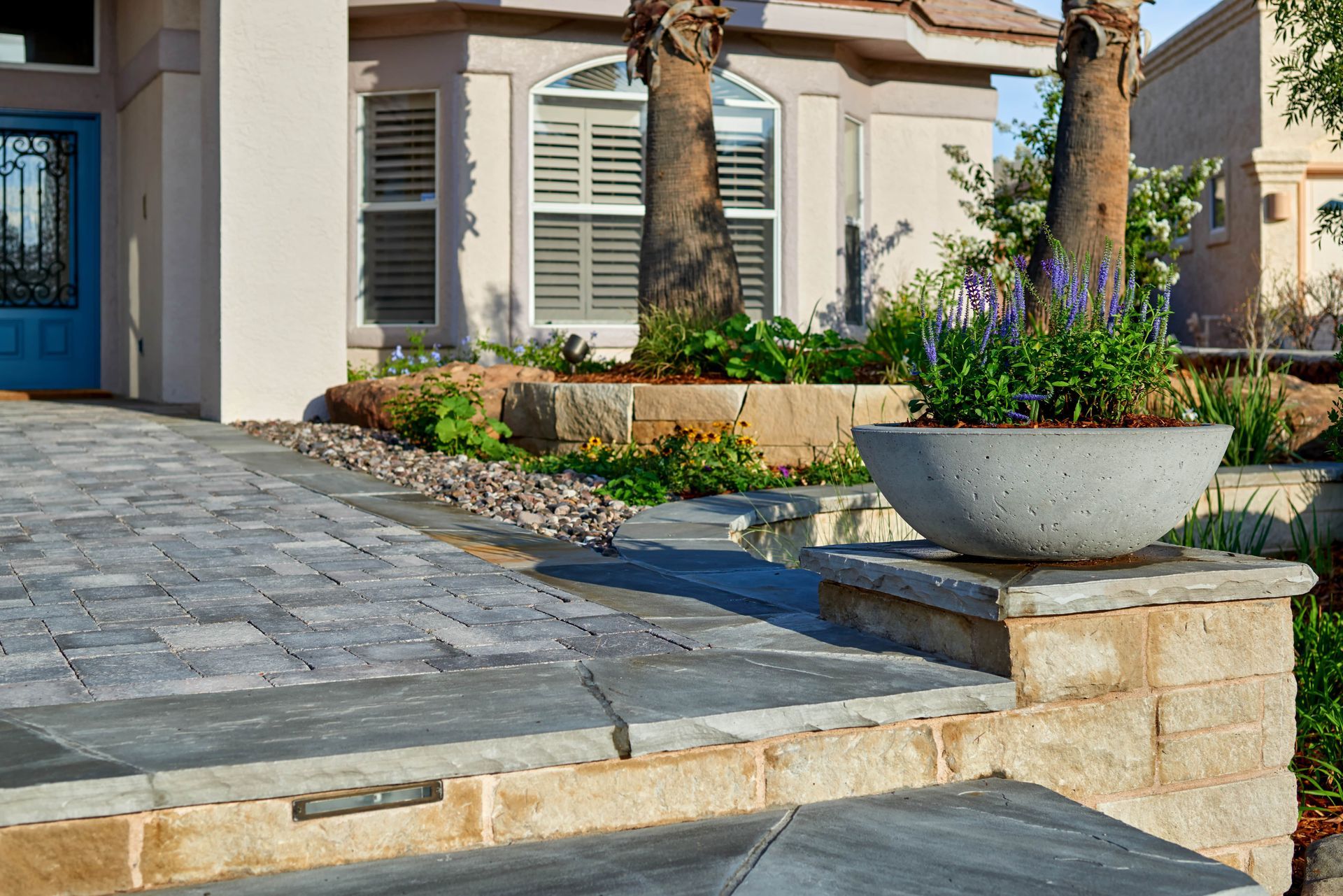 Brick walkway leading to a house with a concrete planter, low retaining wall, and landscaping.