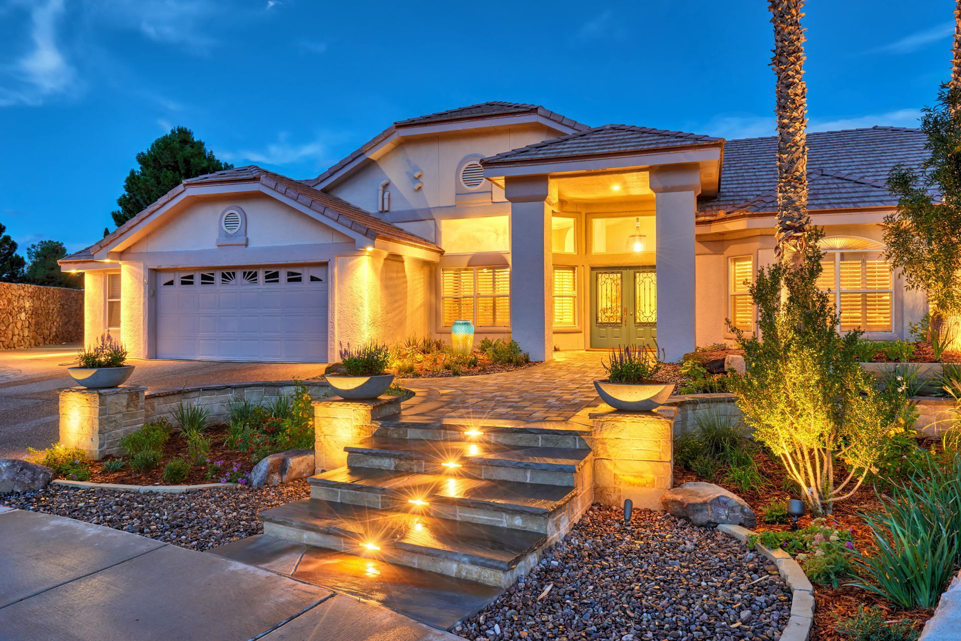 Luxury home exterior with lit steps, landscaping, and a garage at dusk.