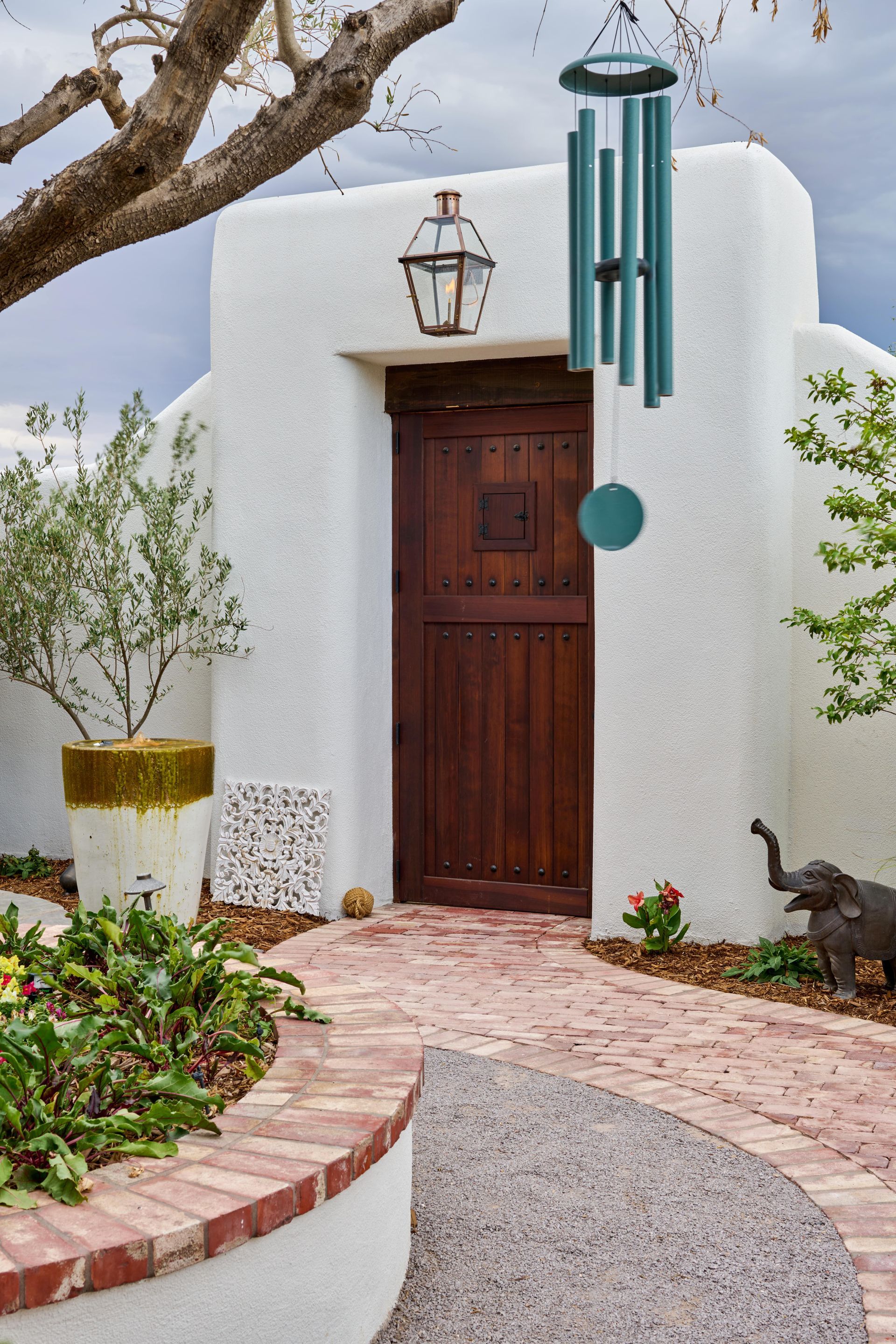 Courtyard entrance with brick path, wooden door, wind chimes, and potted plants.