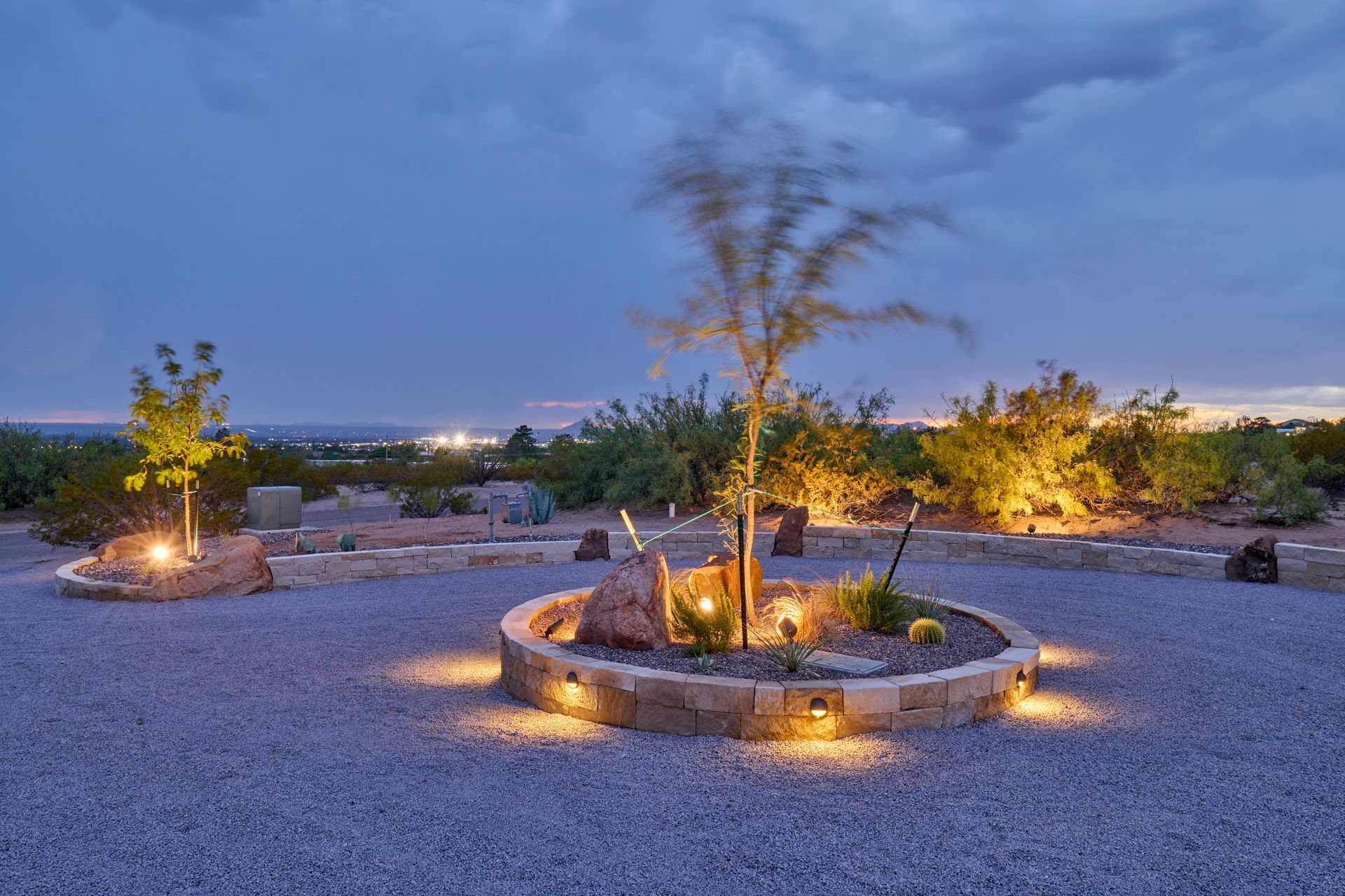 Nighttime outdoor landscape with illuminated trees, rock border, and gravel ground.