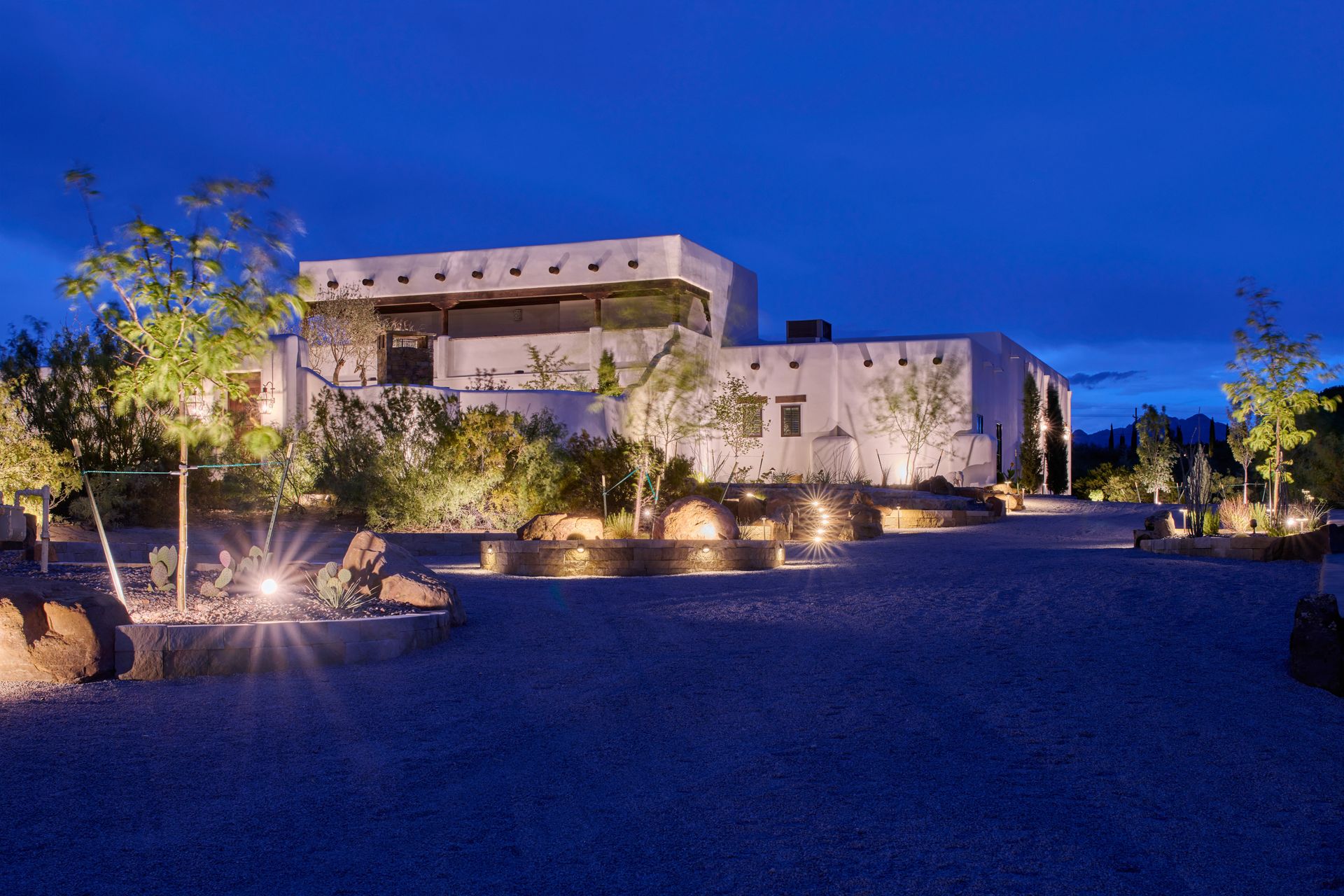 White adobe-style building at night, lit by spotlights, with gravel driveway and sparse desert landscaping.