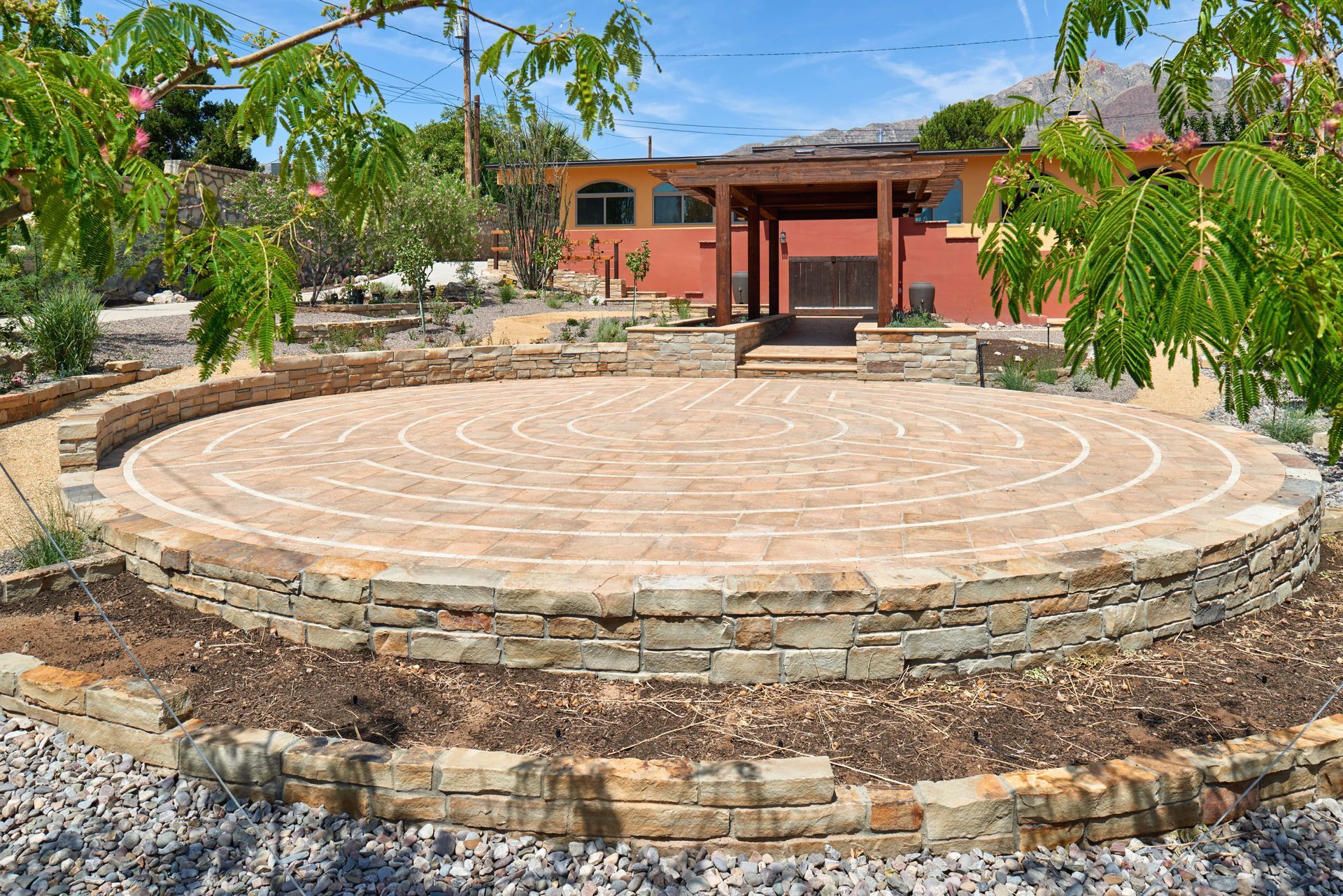 A stone labyrinth with a wooden pergola in the background, set in a garden with trees.