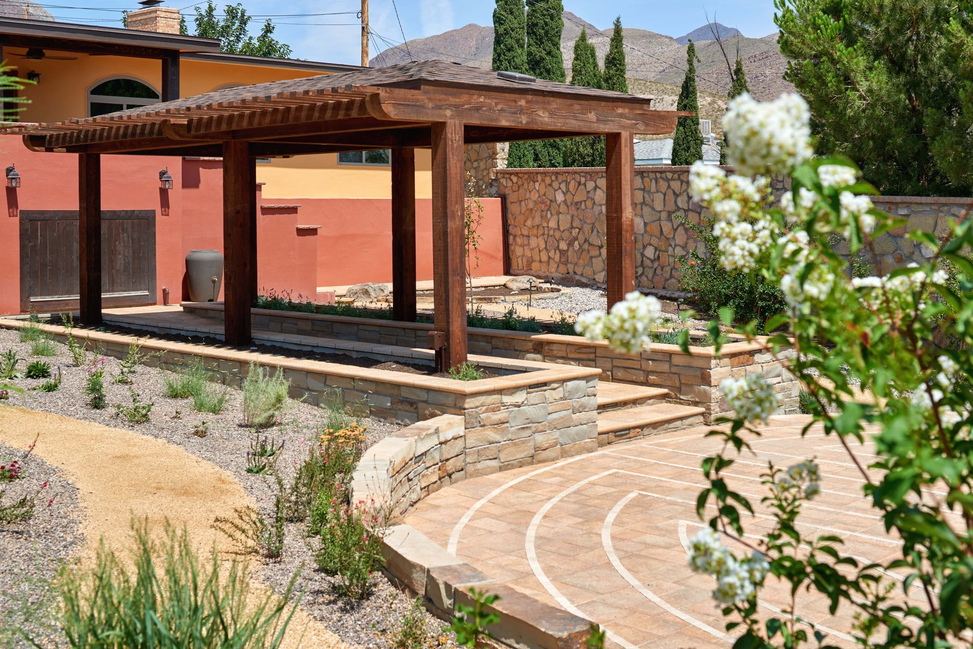 A wooden pergola over a patio with stone steps, path, and tan/orange building. Mountains in background.