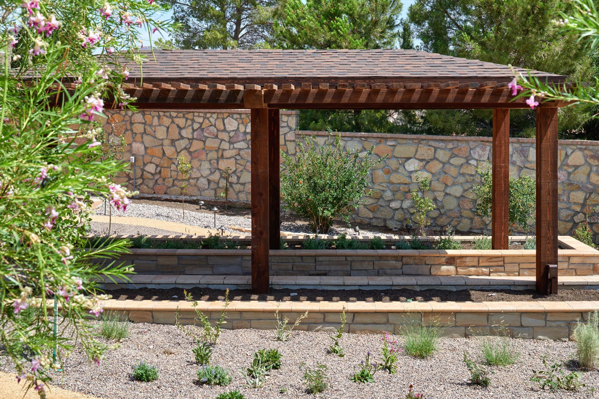 Wooden pergola in a landscaped yard with stone retaining walls and a rock background.