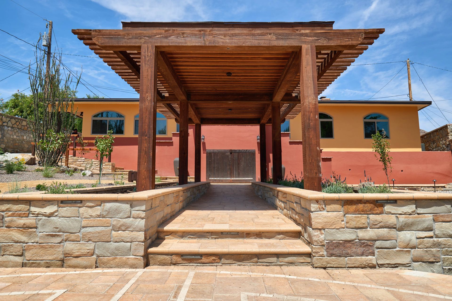 Wooden pergola over a stone walkway leading to a building with orange and yellow walls.