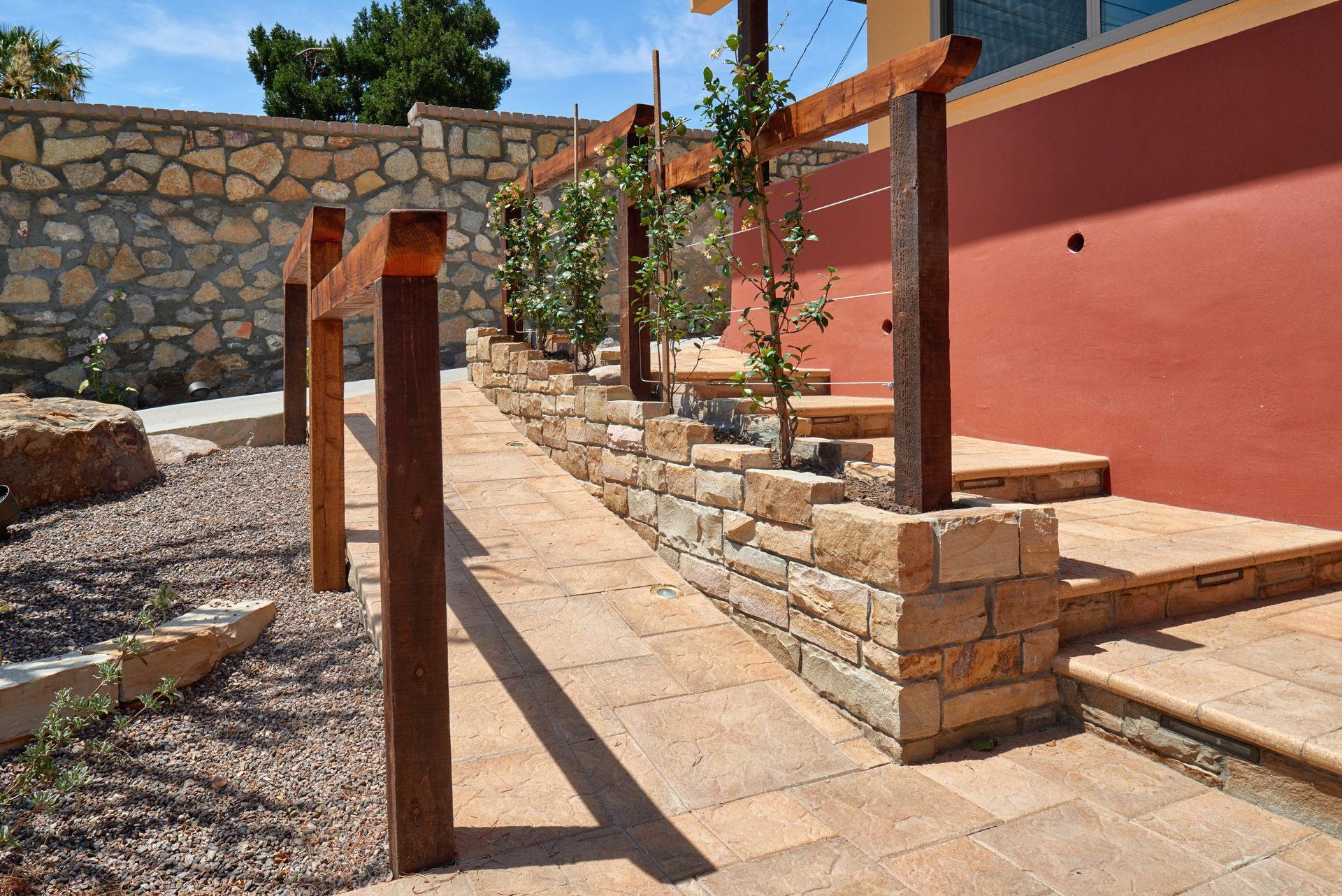 Pathway with stone wall, steps, and wooden trellis. Red building in the background. Sunny outdoor setting.