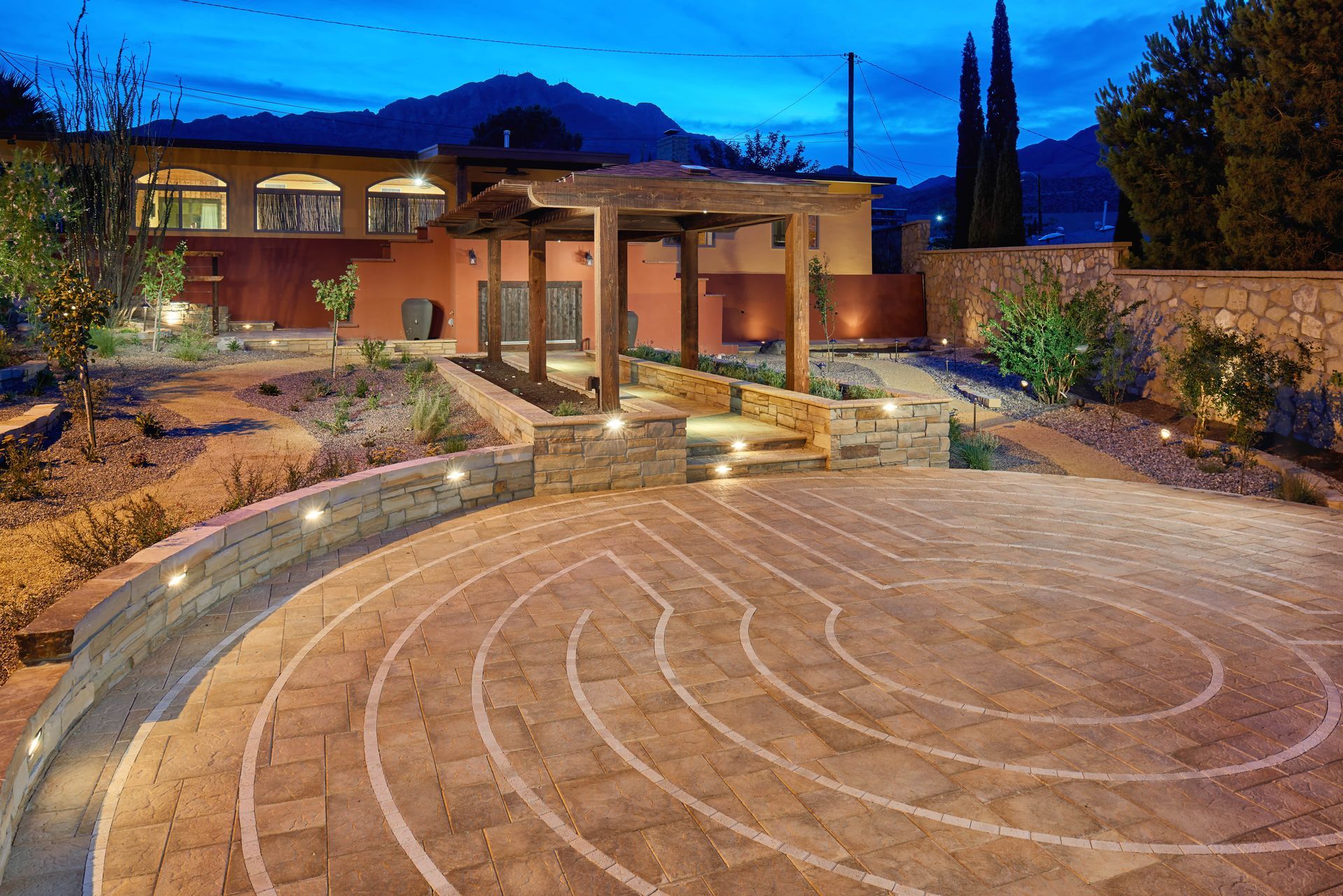 Stone labyrinth with pergola and lit pathway. Building and mountain in the background at dusk.