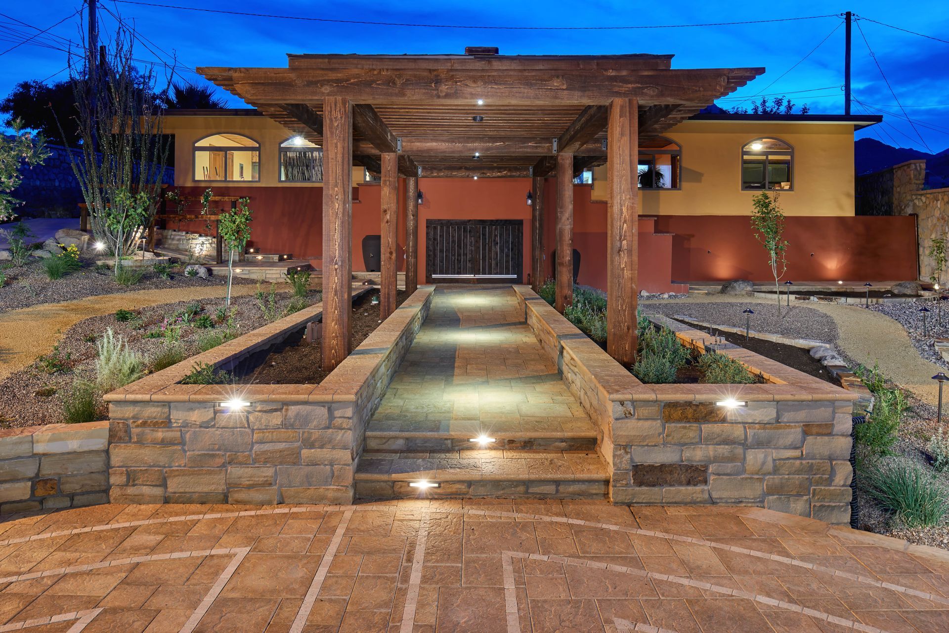 Stone walkway with pergola leading to a home with warm lighting, against a dusk sky.