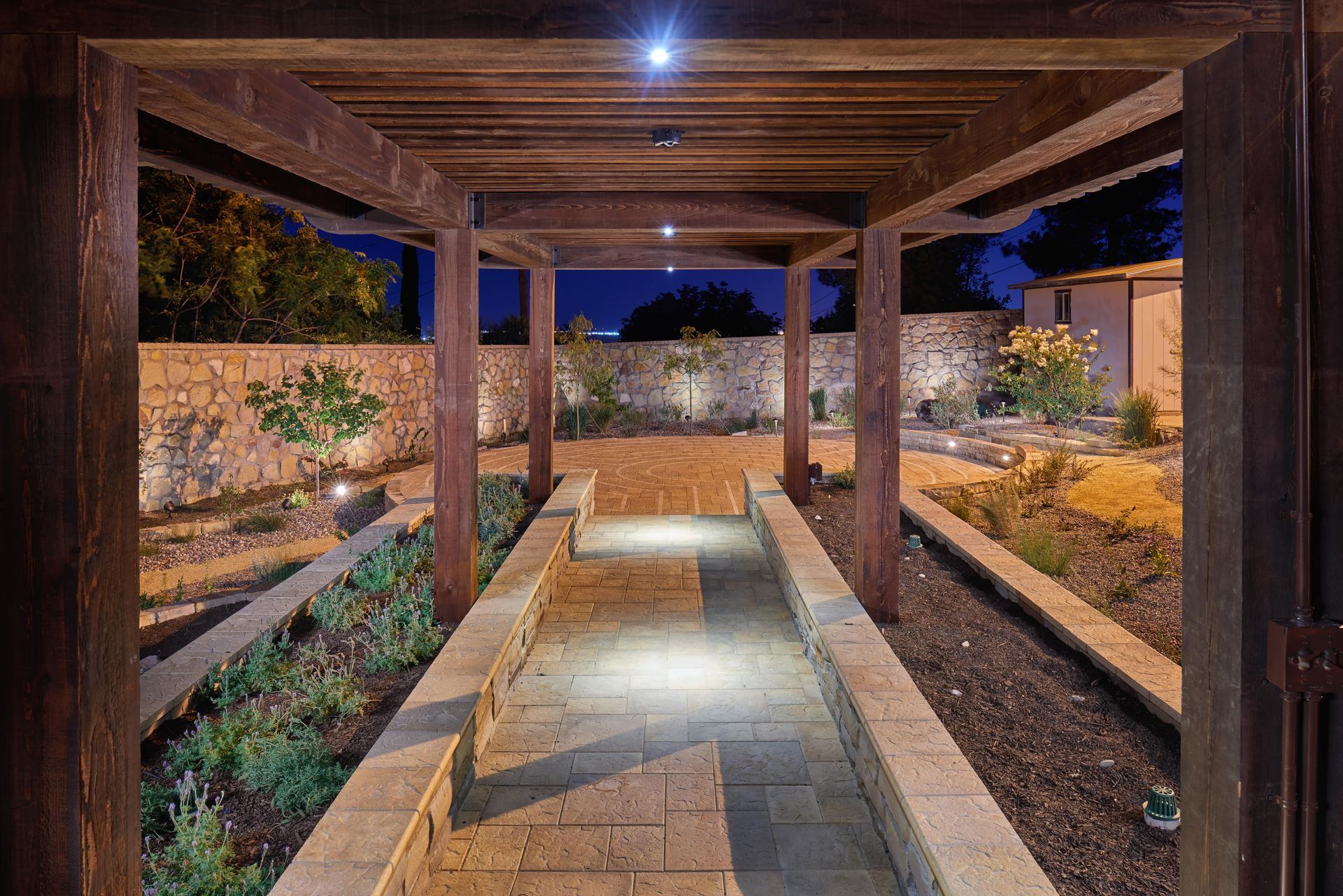 Wooden pergola over a stone path in a garden, lit with overhead and ground lights at night.
