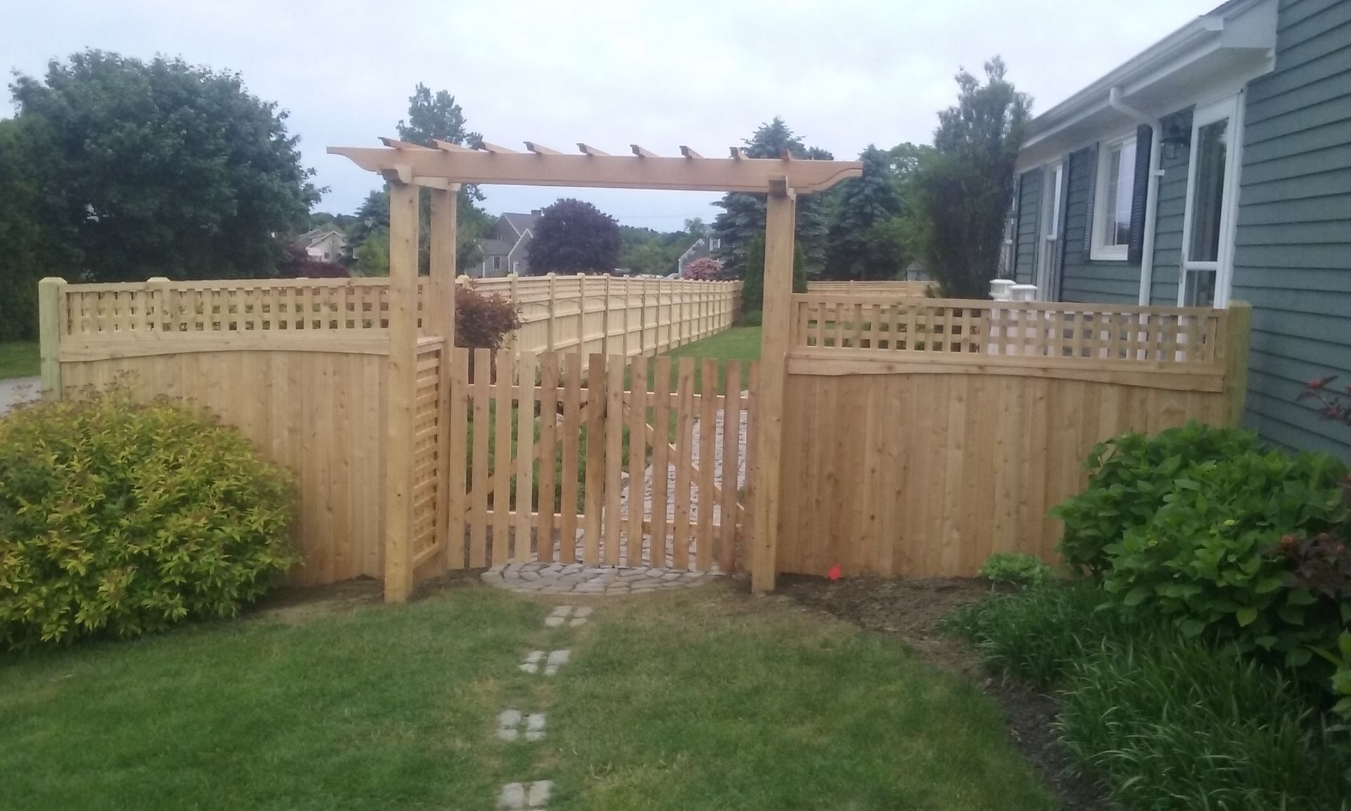 Wooden gate and fence with trellis archway in a yard, leading to a path. Green grass, bushes, and a house.