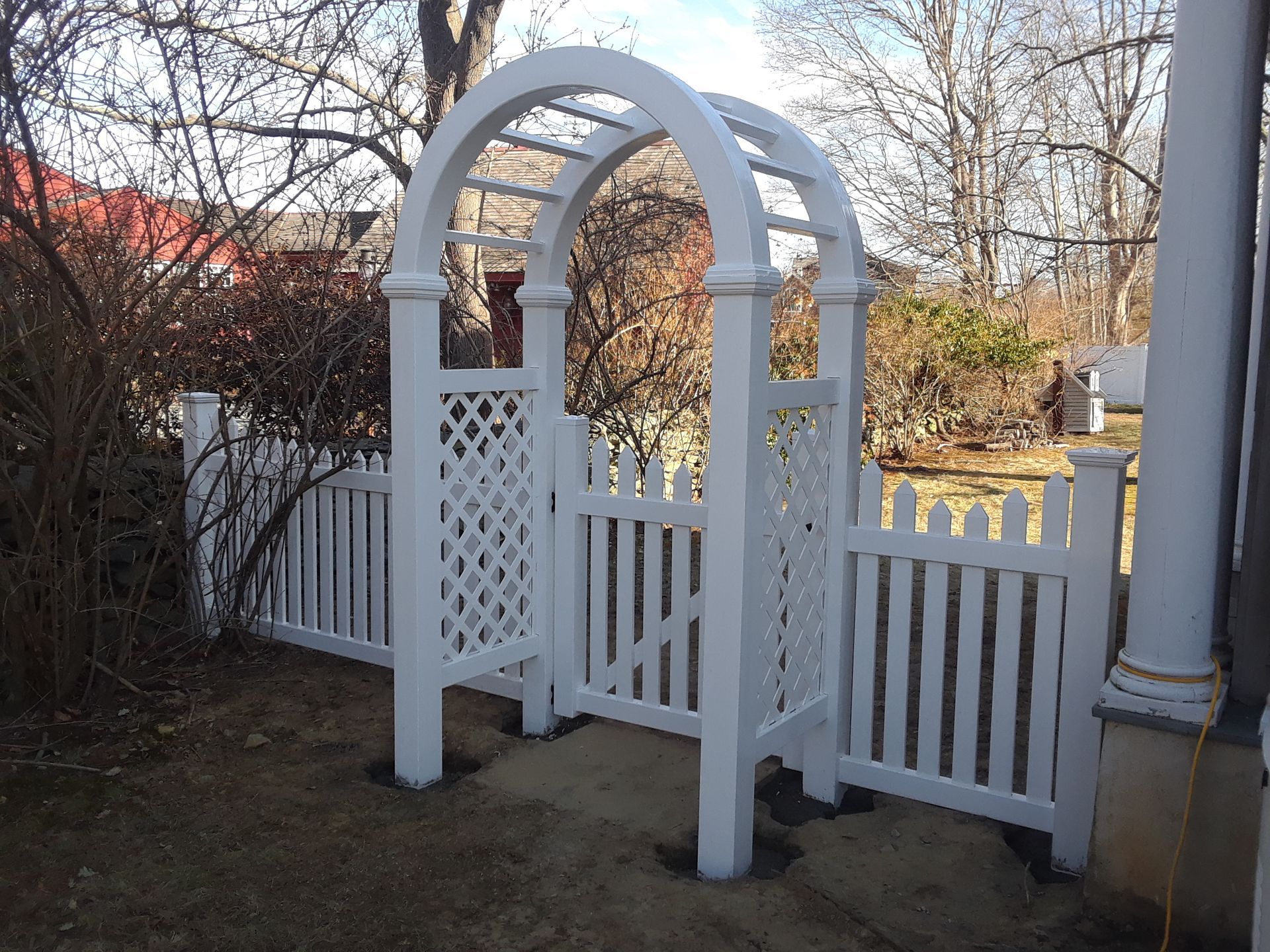 White arched gate with lattice panels, surrounded by a white picket fence, on a sunny day.