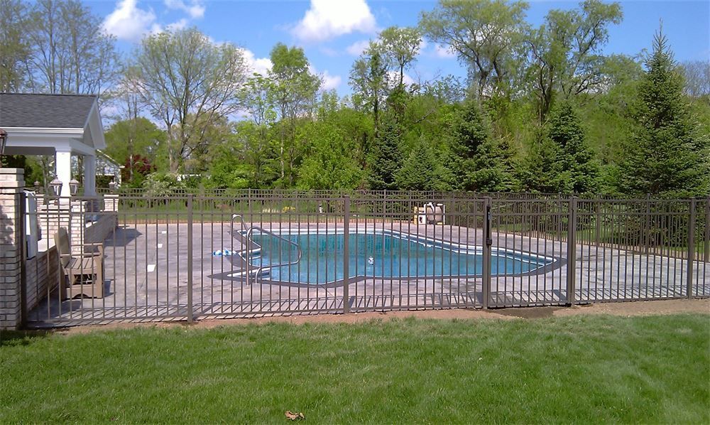 Pool surrounded by a dark metal fence, behind a green lawn. Trees and a house are visible in the background.