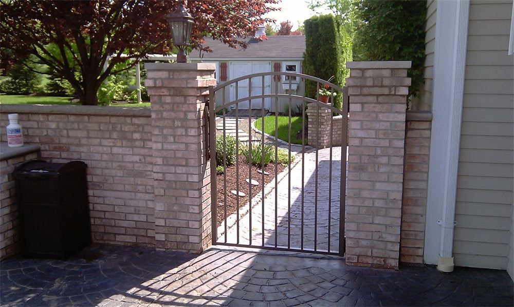 Brick wall with wrought-iron gate between brick pillars; leads to a path. Trash can and house present.