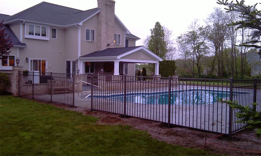 A backyard pool enclosed by a black metal fence, next to a large two-story house with a covered patio.