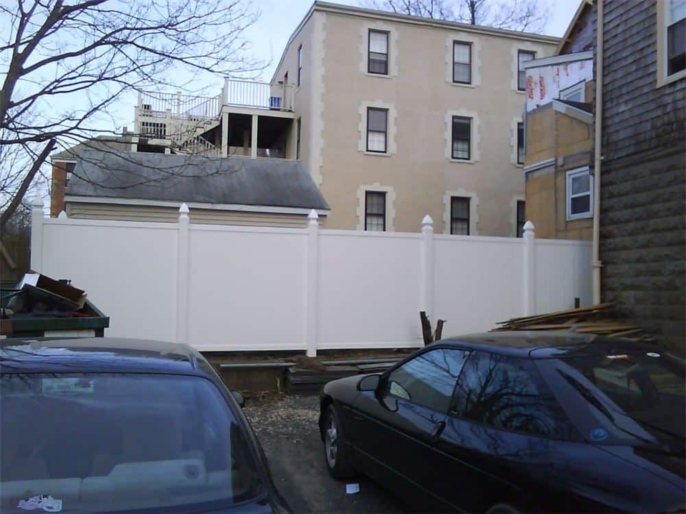 White vinyl fence in front of a multi-story building and parked cars.