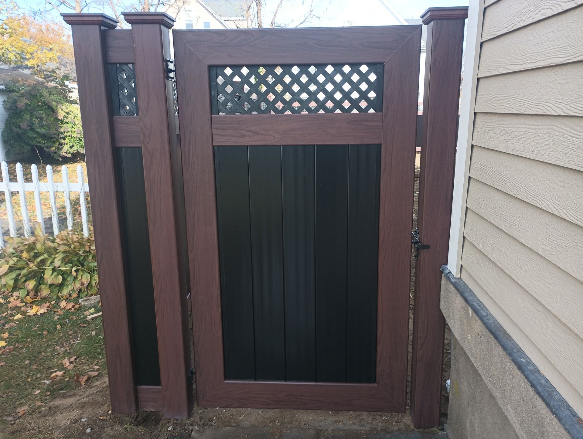 Brown and black wooden gate with lattice top section and vertical paneling, adjacent to a house.