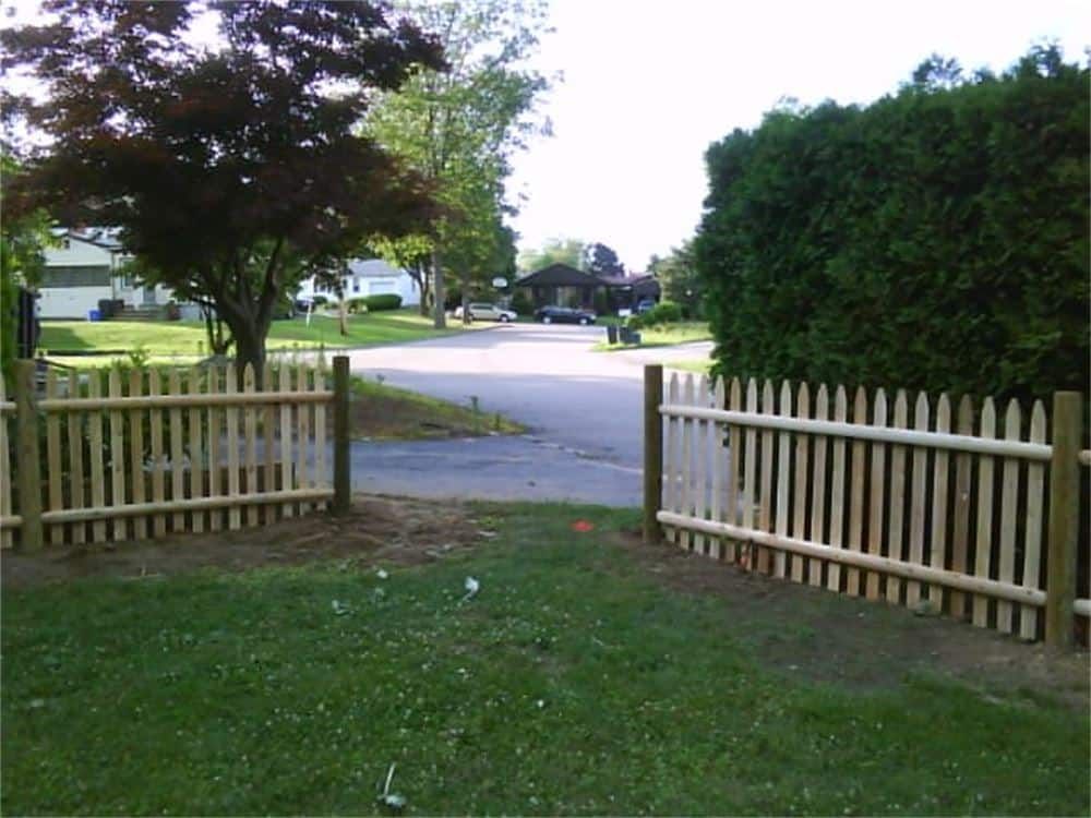 Wooden picket fence with open gate leading to a residential street with houses and trees.