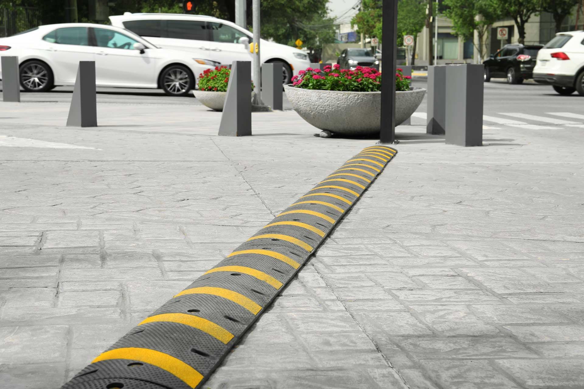 Speed bump on a gray brick sidewalk near a crosswalk with cars and flower planters in the background.