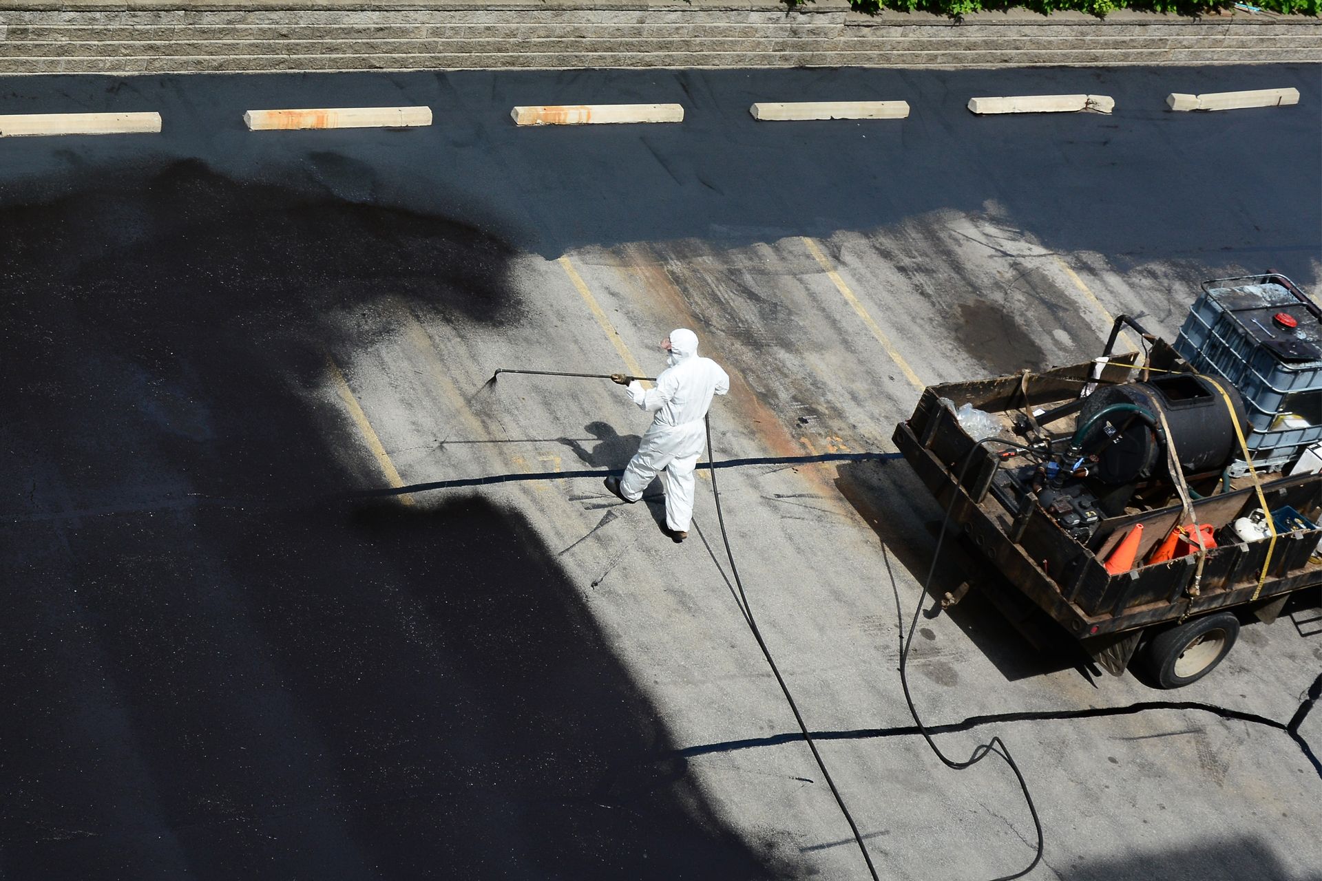 Person in white protective suit sealing cracks in asphalt with equipment in a parking lot.