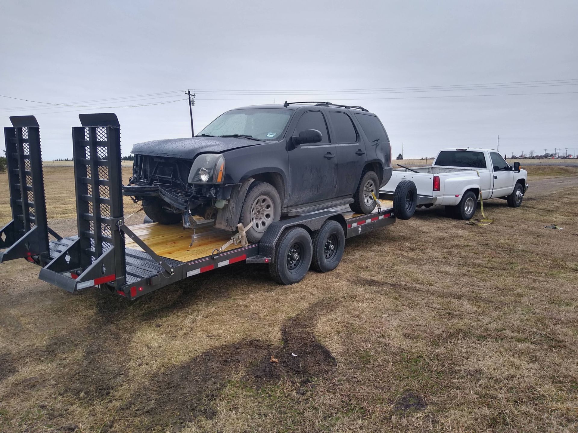 A black damaged SUV loaded on a trailer being towed by a white pickup truck.