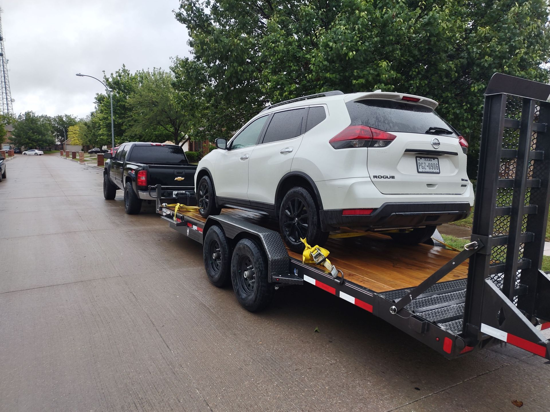 Black pickup truck towing a white SUV on a trailer down a wet street.