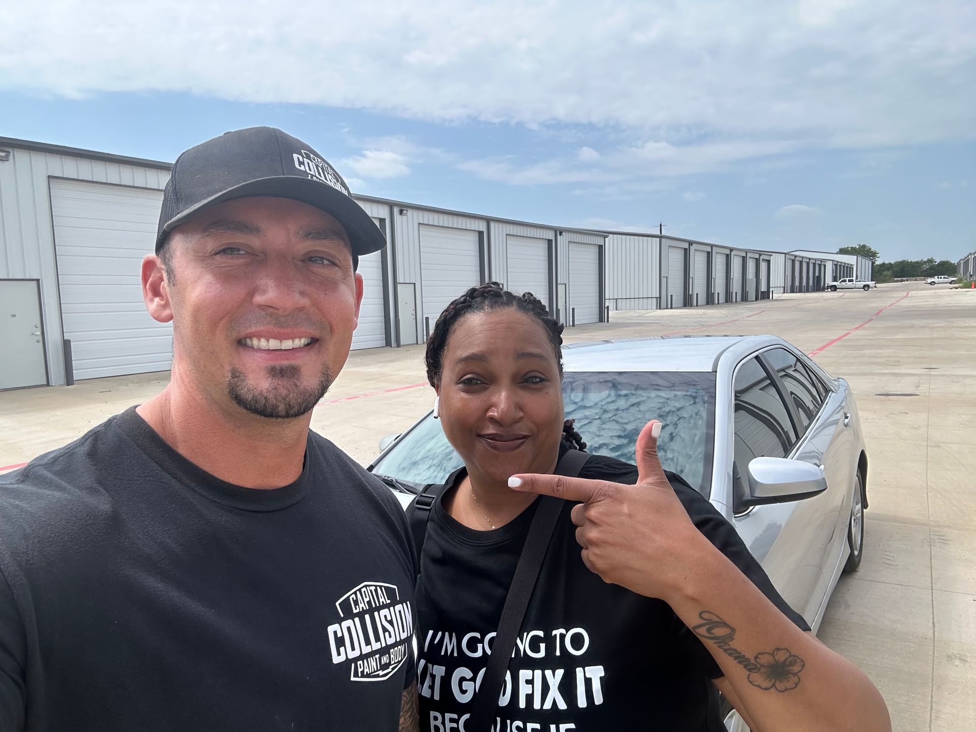 Two people smile near a silver car in front of storage units. One points.