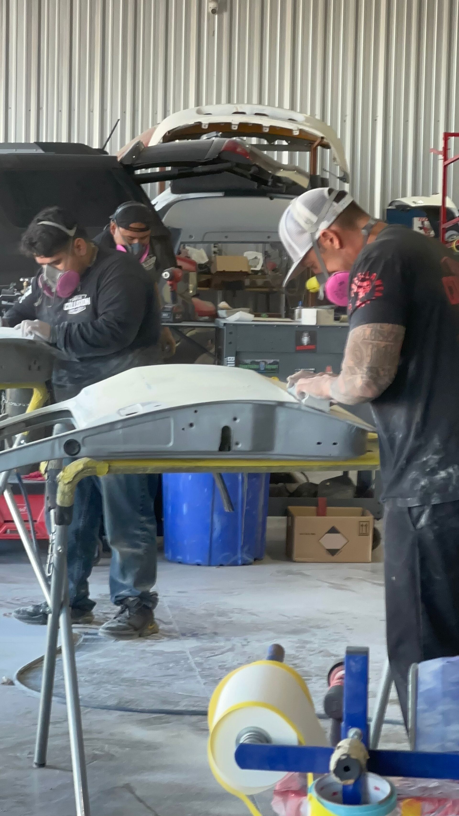 Three people sanding car body parts in a workshop, wearing masks for protection.