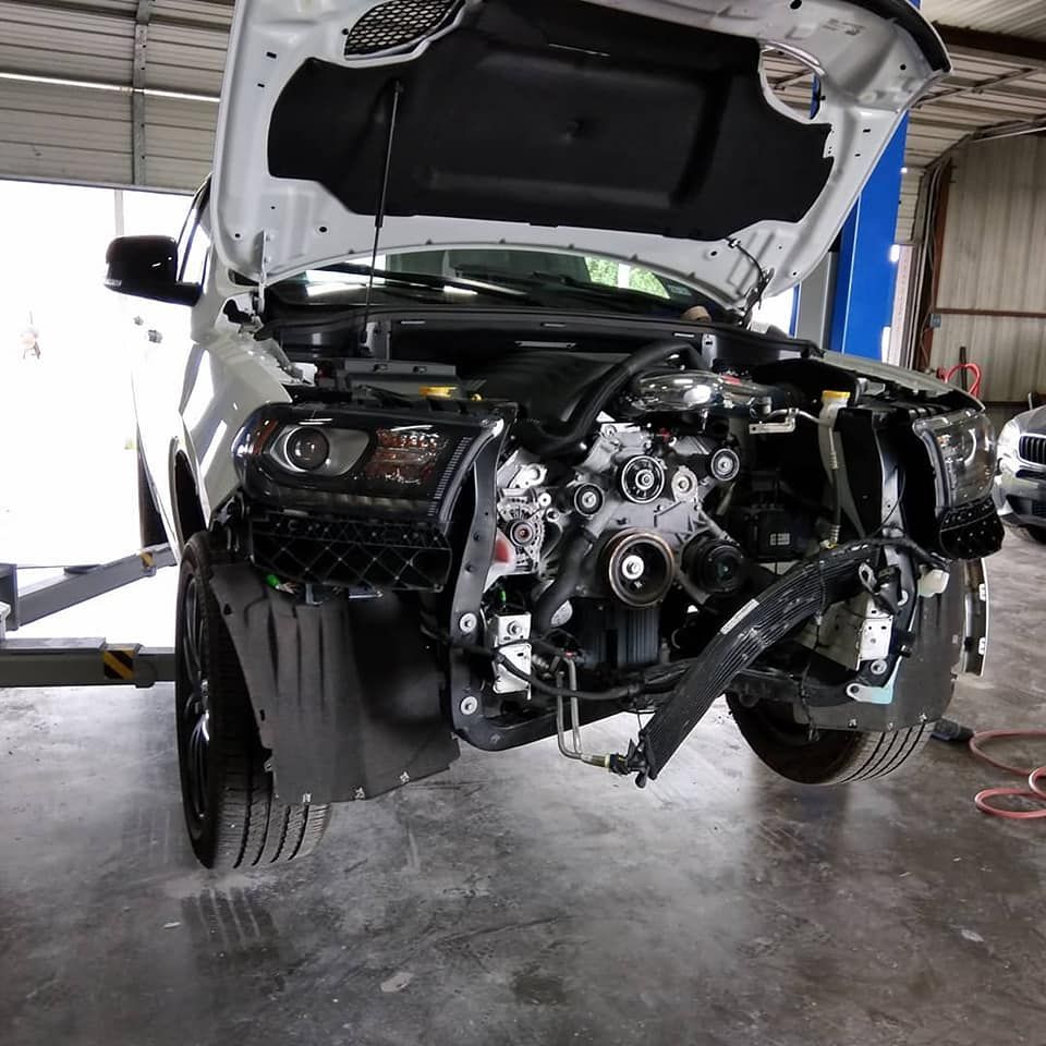 White pickup truck with front-end damage, hood open, in a repair shop.