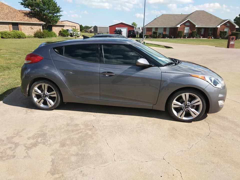 Gray Hyundai Veloster car parked on concrete driveway in front of houses.