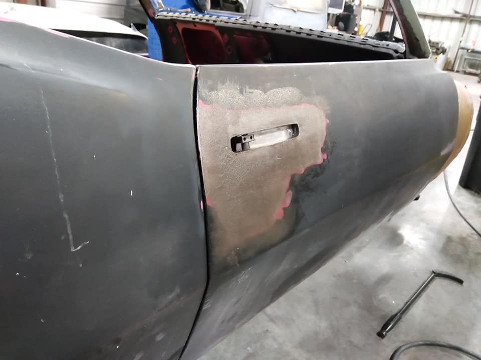 Car door in a workshop, dark grey with a patched section, door handle slot, next to another panel.