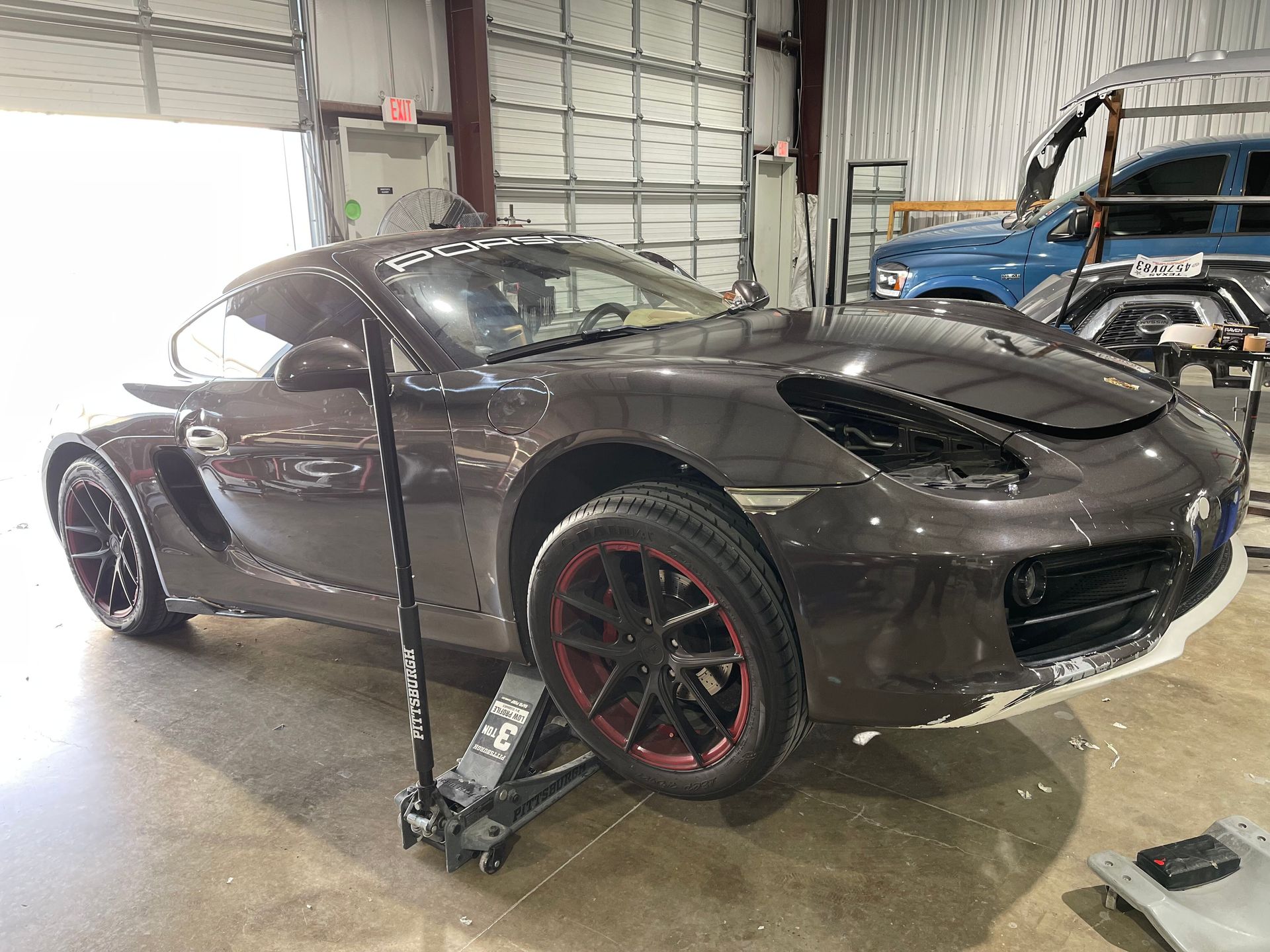 A brown sports car on a jack in a garage, undergoing repair. Black and red rims are visible.