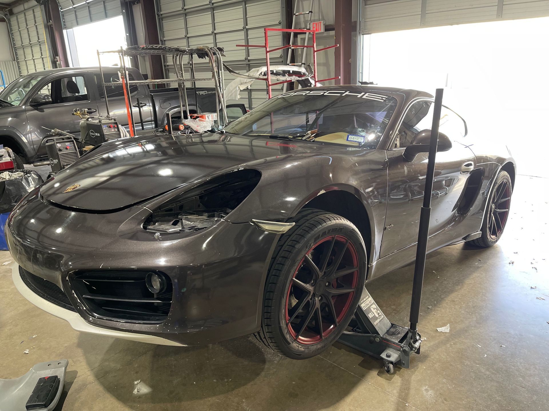 A grey Porsche Cayman being worked on in a garage; front bumper removed, jacked up, and wheels with red accents.
