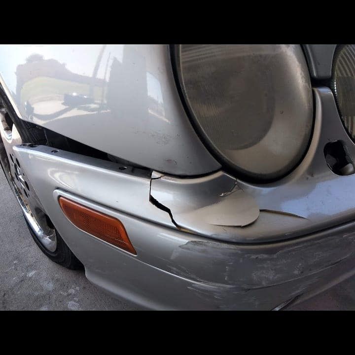Damaged silver car bumper with a cracked area near the headlight and turn signal.