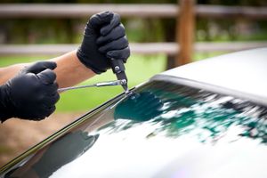 Hands in black gloves using a tool to cut a black strip on a car windshield.