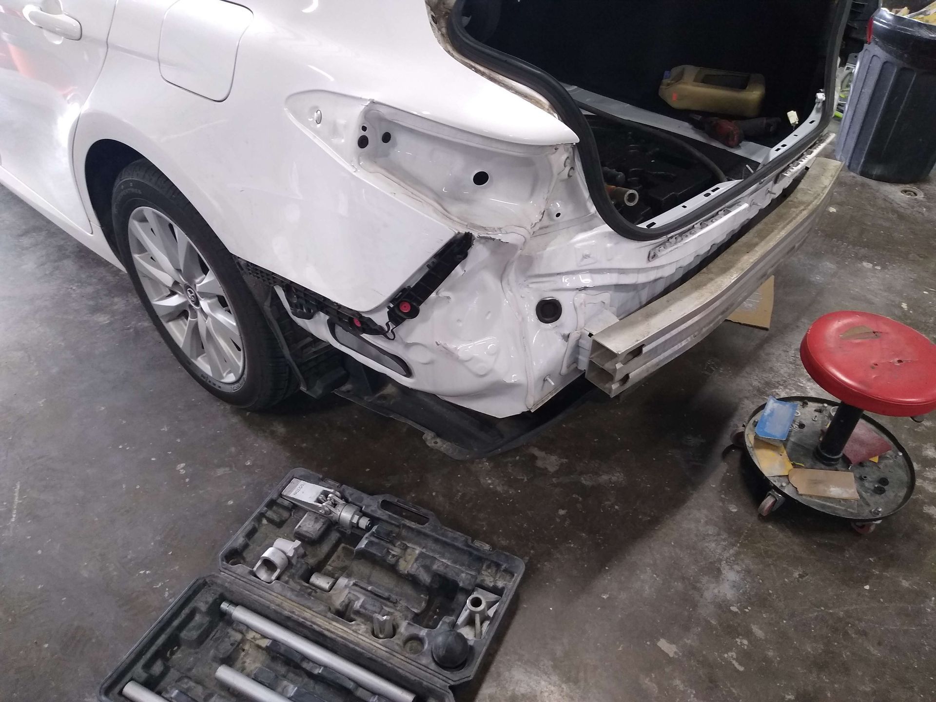 White car with rear bumper removed, revealing damage. Tools and red stool in a garage setting.