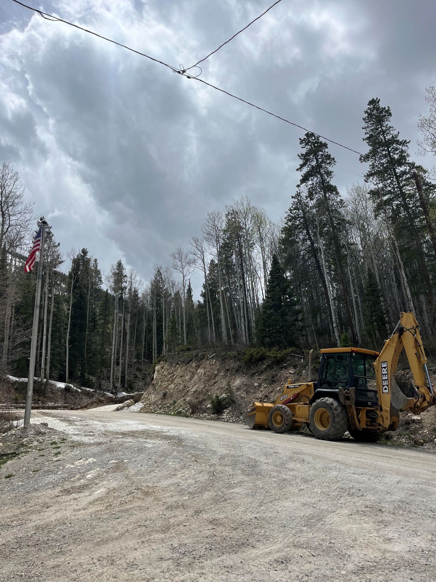 Construction site with yellow excavator on a dirt road beside a forested hillside under cloudy skies