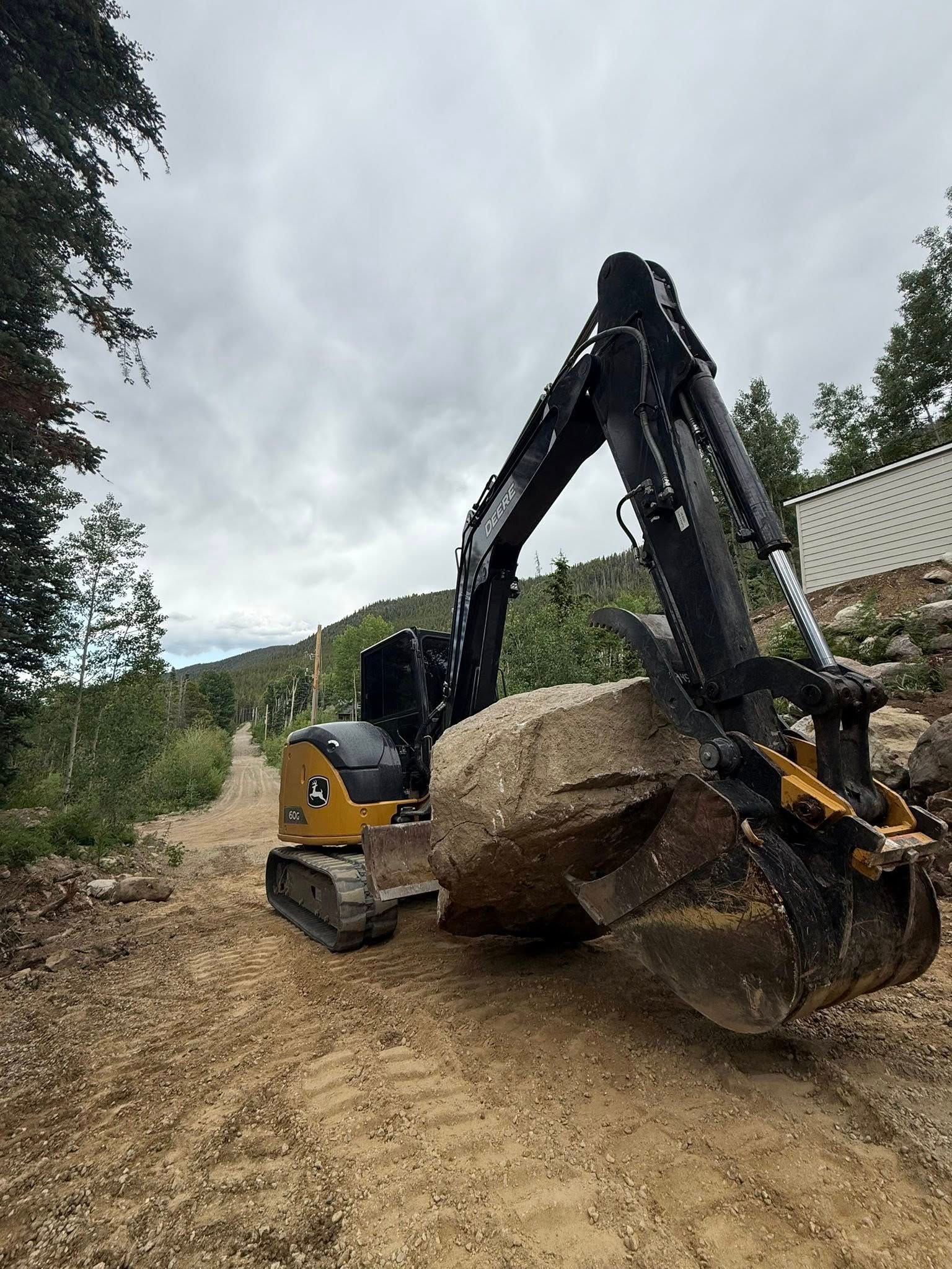 Yellow excavator lifting a large boulder on a dirt road under an overcast sky