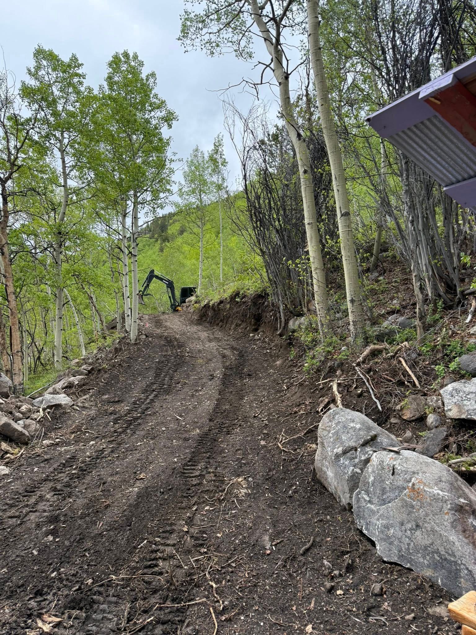 Steep dirt trail through a forest, with rocks on the right and a building edge at the top right.