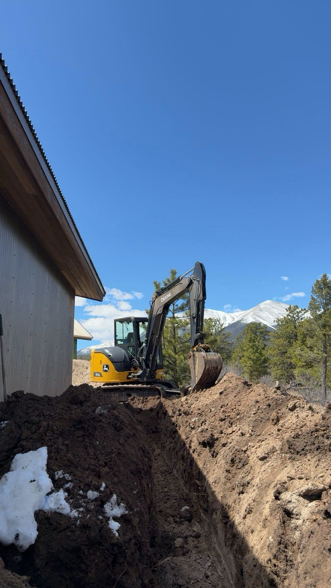 Excavator digging a trench beside a white building, with snow, trees, and mountains under a clear blue sky.