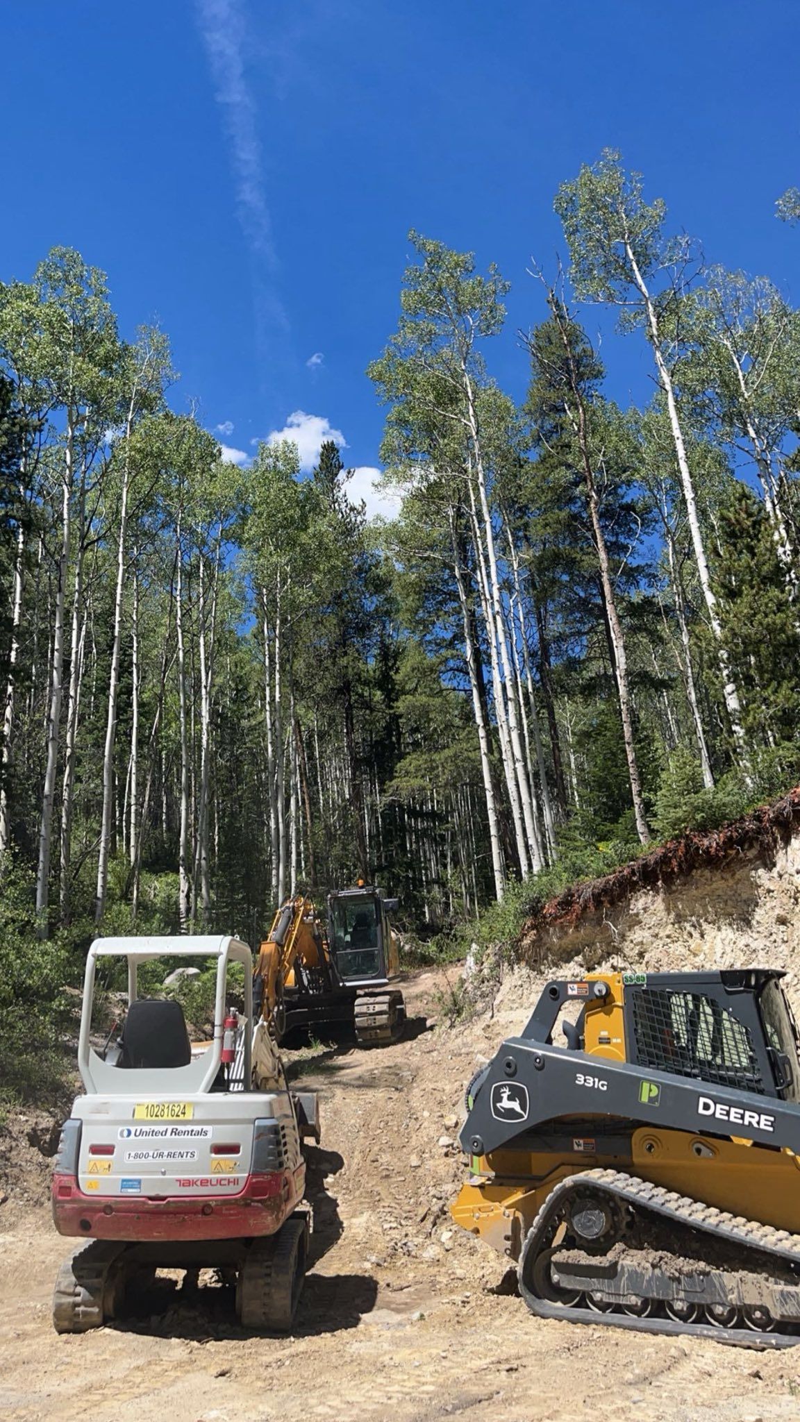Two off-road vehicles parked on a dirt slope in a forest clearing under a blue sky.