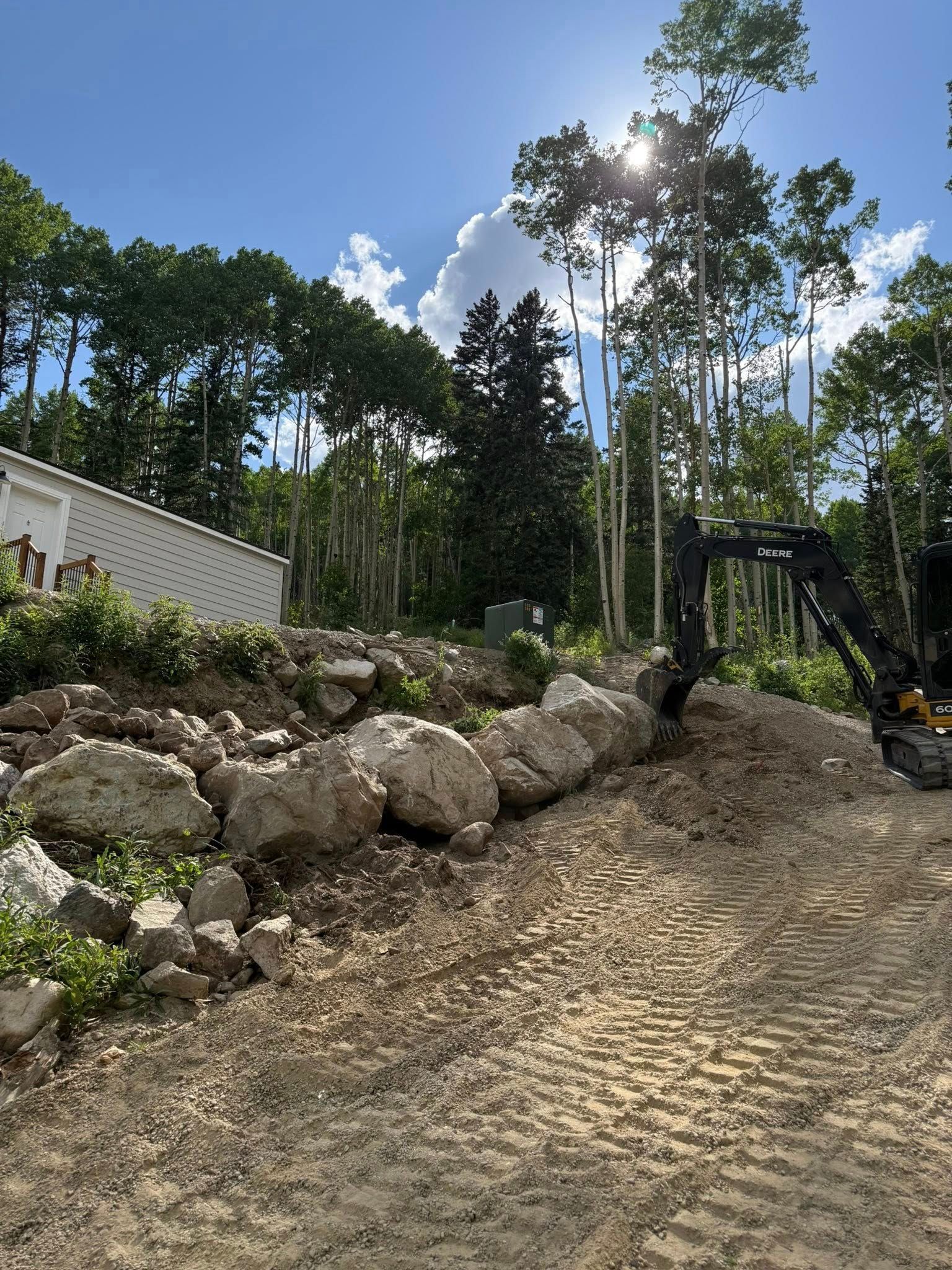 Rock-lined dirt path on a sunny hillside with tall trees and a white house to the left.
